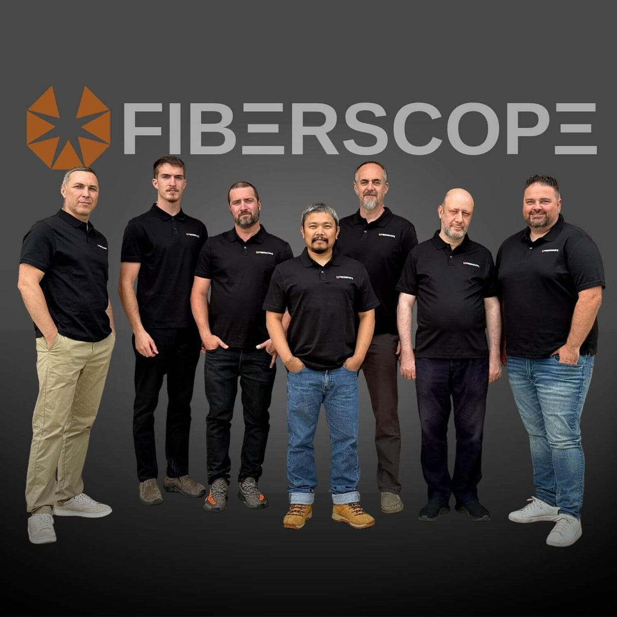 The team of FIBERSCOPE standing in front of a dark gray background with the FIBERSCOPE logo and name. They are wearing matching black polo shirts with a small white logo on the chest, paired with various pants in beige, black, brown, and blue jeans.
