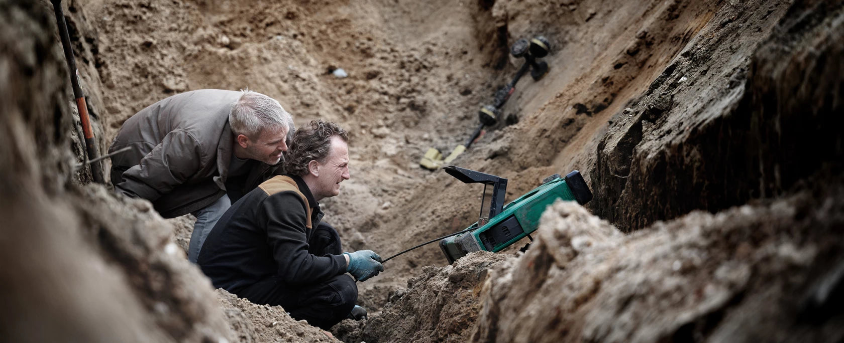Two contractors crouched in a dirt excavation trench, with one using a handheld MiniFlex drain inspection camera and the other observing closely.
