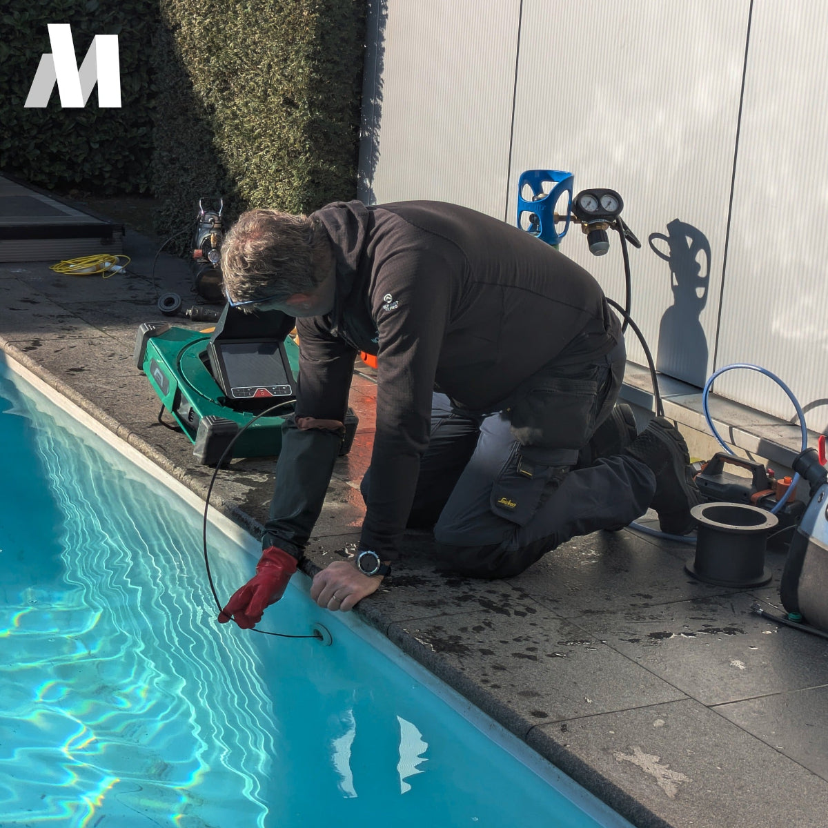 A pool maintenance worker kneels by a swimming pool, using a MiniFlex small pipe camera in the water while wearing a dark uniform and red gloves. Pool equipment, hoses, and a white wall are visible in the background.