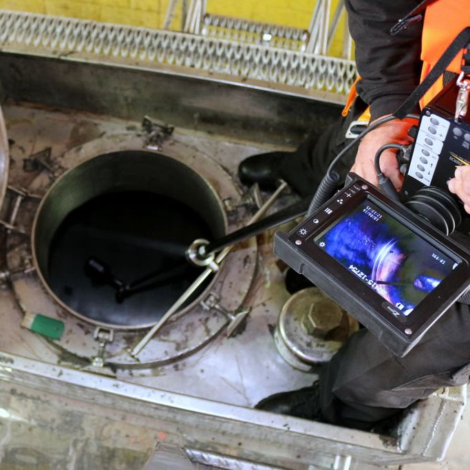 A close-up action shot of a professional inspection worker using a pole-mounted inspection camera inside a tank. The worker holds a handheld monitor displaying a live camera feed, which is positioned inside a tanker metal access pit with its lid swung open to the left.
